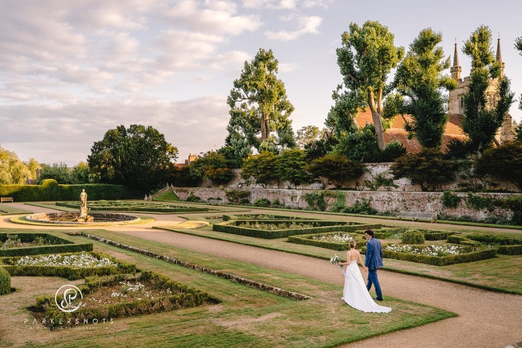Wedding Photograph from Penshurst Place from Anna & Harry's wedding by Parkershots. Image shows couple walking in Rose Garden near sunset with the church behind them